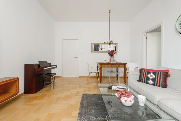 A living room with a piano, a dining table with chairs, a sofa with a patterned cushion, and a coffee table with flowers and a mug.