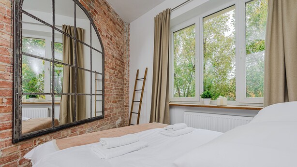 A room with a bed, a large mirror, a brick wall, a window with curtains, and a radiator. - P&O Apartments Suligowskiego (Warsaw)