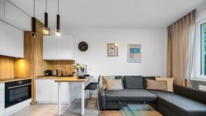 Una sala de estar moderna con un sofá gris, una mesa de centro de vidrio y una zona de cocina con armarios blancos y una encimera de madera.