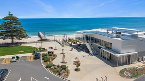 Een modern gebouw met een glazen gevel, met uitzicht op een strand, met terrasplekken en een trap naar de ingang.