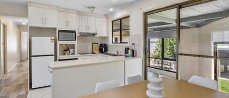 A modern kitchen with white cabinetry, a built-in microwave, and a coffee maker. There is a dining area with a wooden table and chairs, and a large window offering a view of the outdoors.
