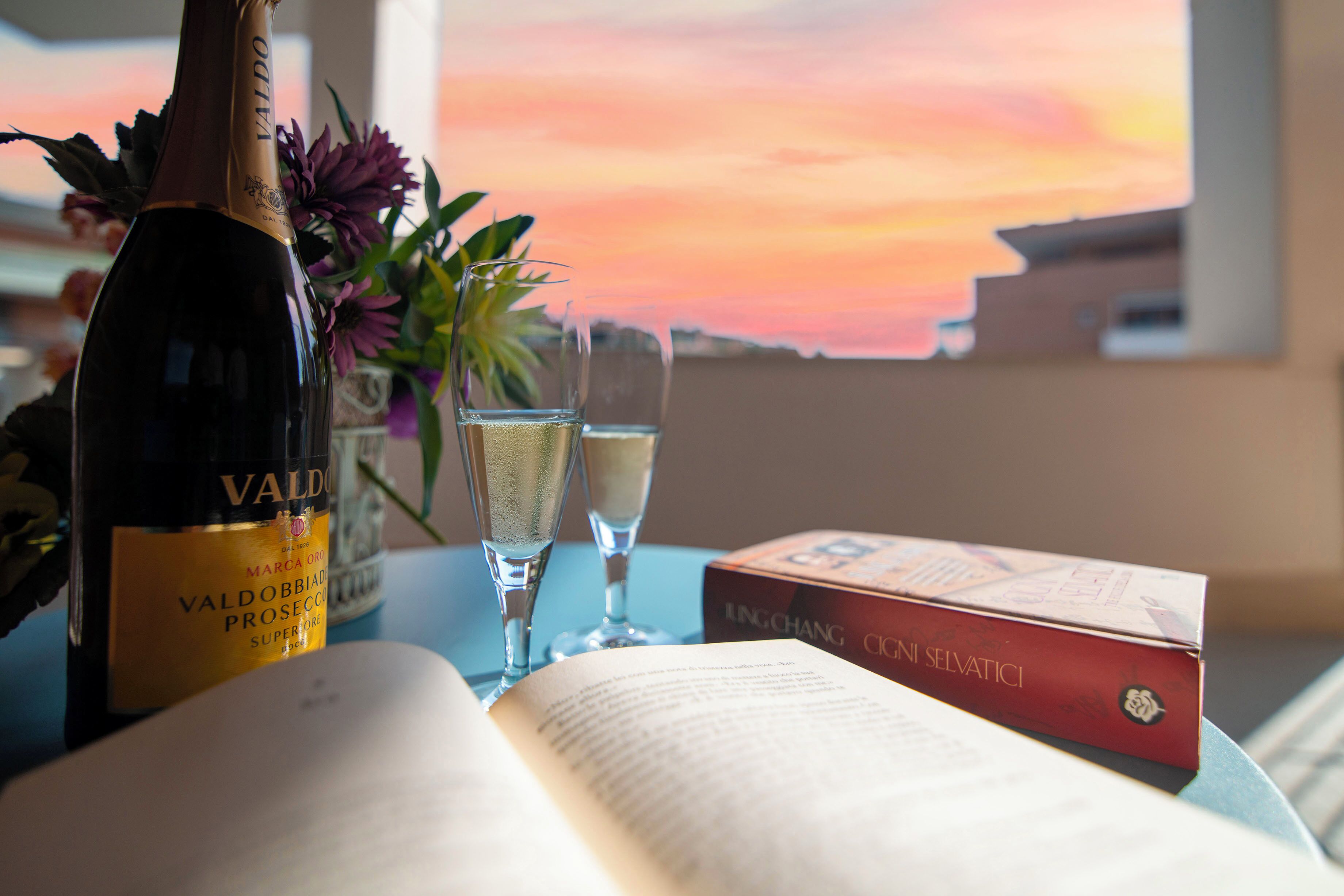 A bottle of Valdobbiadri Prosecco, two champagne flutes, a book, and a box of cigars on a table with a view of a sunset.