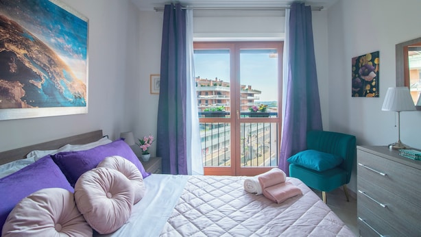 A bedroom with a bed, a chair, a dresser, and a balcony with a view of buildings.