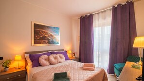 A neatly made bed with a quilt, pillows, and folded towels. There is a bedside table with a lamp and a small plant. A painting of a landscape hangs on the wall.