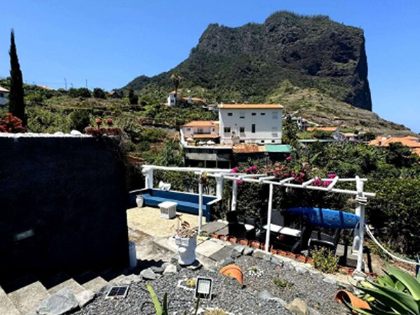 A coastal view with a mountain in the background, a white building with a red roof, and a garden area with plants and a blue structure.