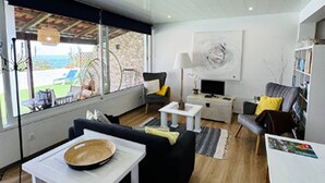A modern living room with a sofa, coffee table, and TV stand. There is a large window offering a view of the outdoors. - Studio in Porto da Cruz With Ocean Views (Machico)