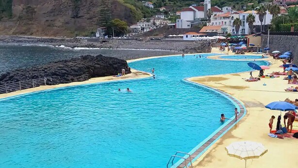 A coastal swimming pool with multiple lanes, a sandy beach area with umbrellas, and a backdrop of houses and cliffs. - Studio in Porto da Cruz With Ocean Views (Machico)