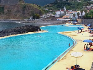 A coastal swimming pool with multiple lanes, a sandy beach area with umbrellas, and a backdrop of houses and cliffs.