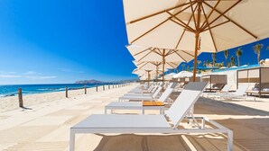 Un hotel frente a la playa con tumbonas y sombrillas, un cielo azul despejado y una playa de arena.