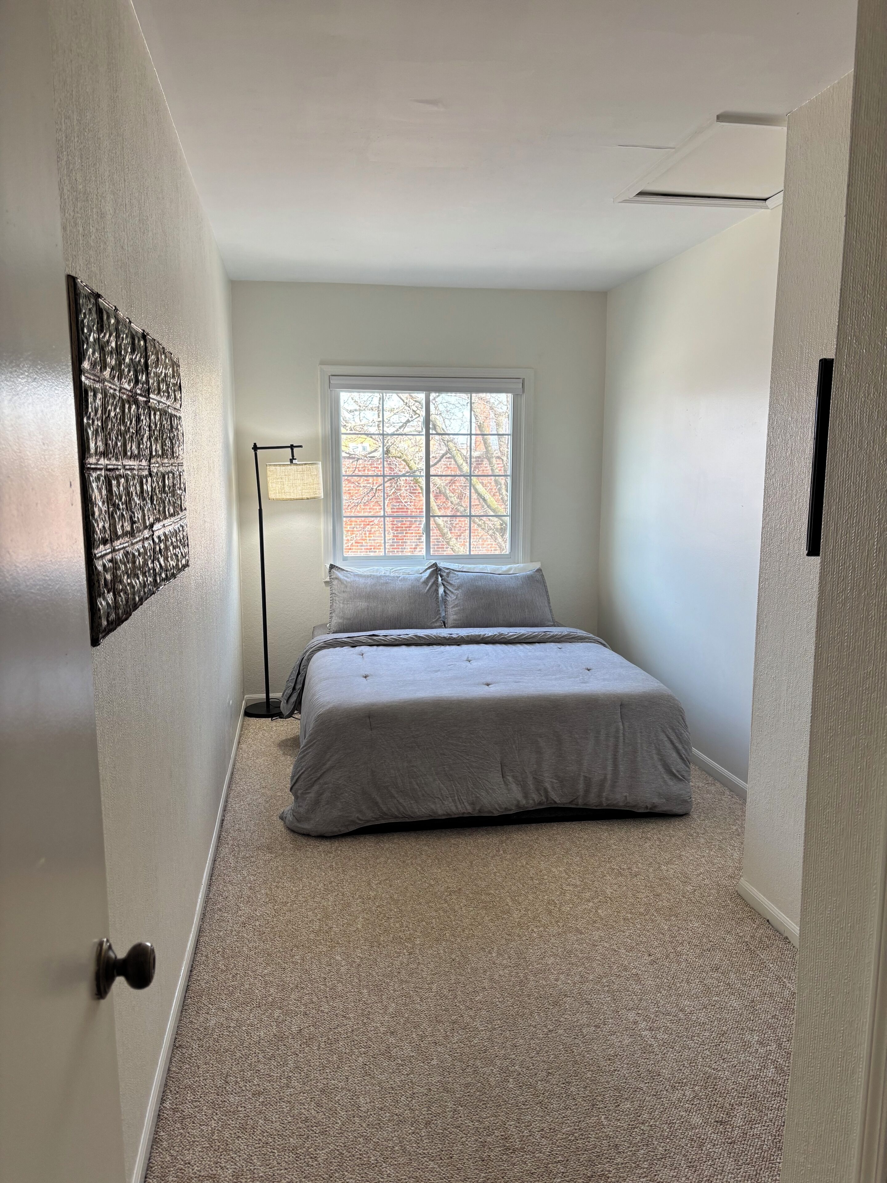A bedroom with a large window, a bed with grey bedding, a floor lamp, and a textured wall decoration.