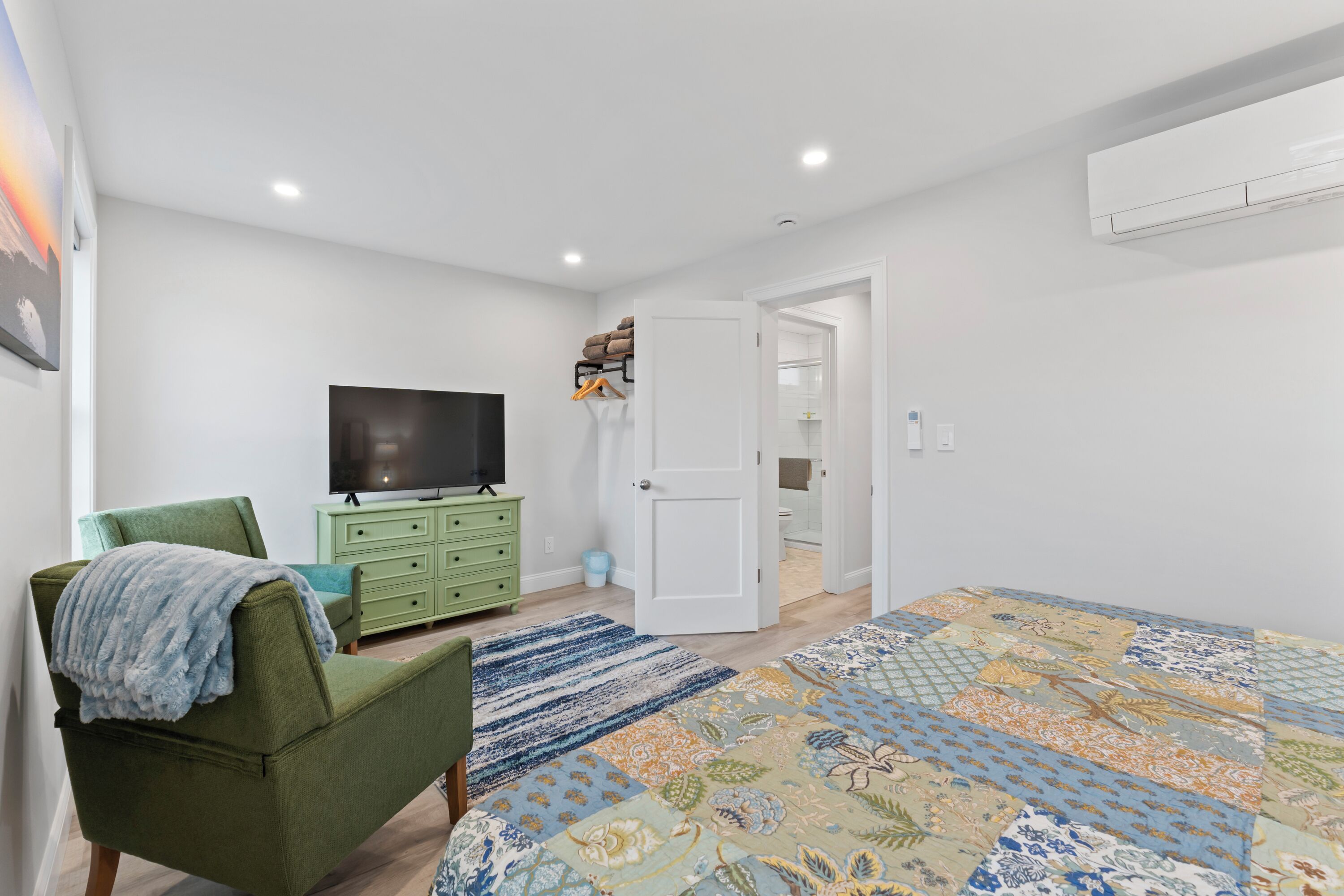 A bedroom with a green armchair, a television, a green dresser, a patterned bedspread, and a door leading to a bathroom.