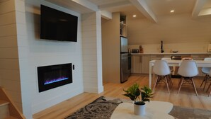 A modern living room with a fireplace, a flat-screen TV, and a dining area with white chairs and a table. - A-Frame retreat with hot tub, sauna (Huntsville)