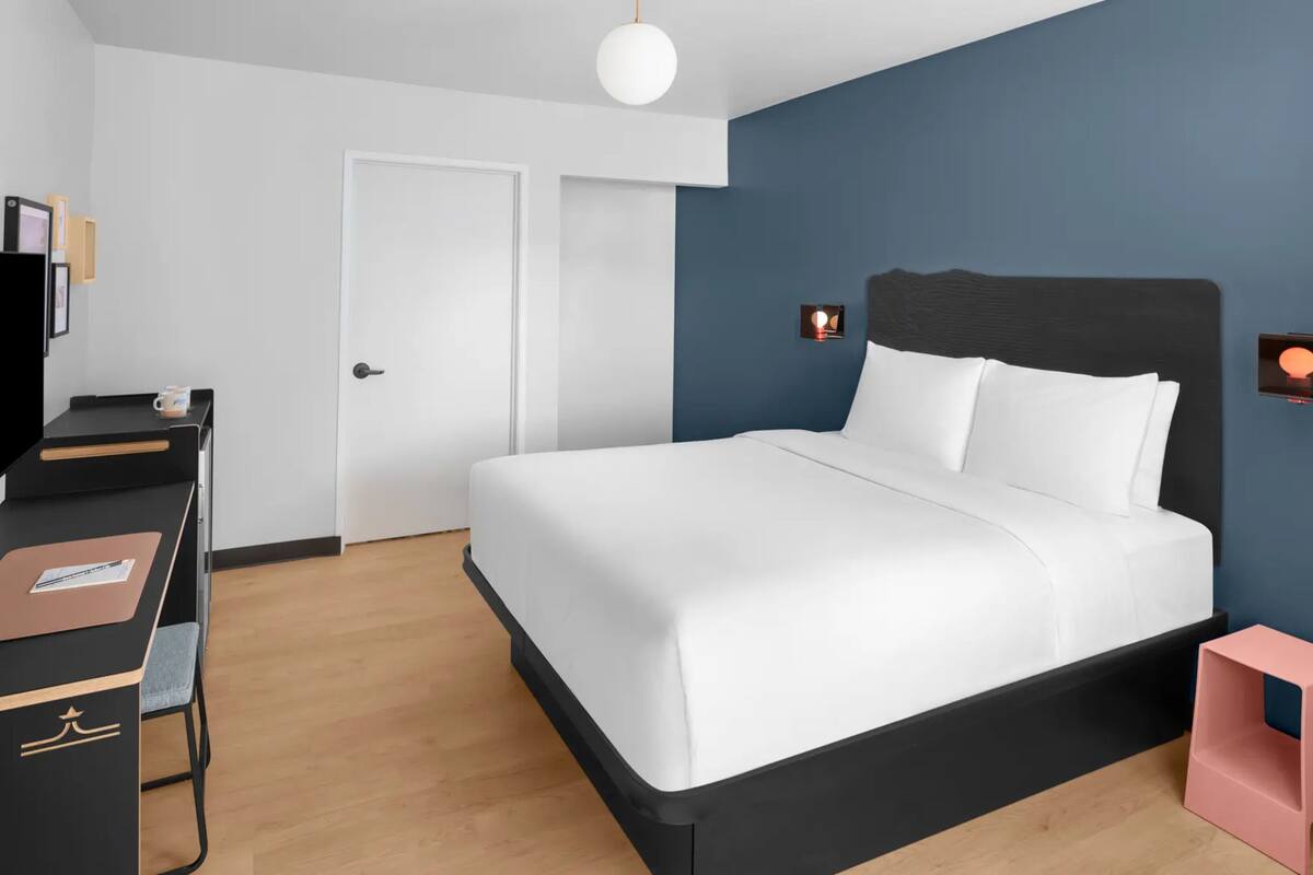 A modern hotel room with a bed, a desk, a television, and a small pink stool.