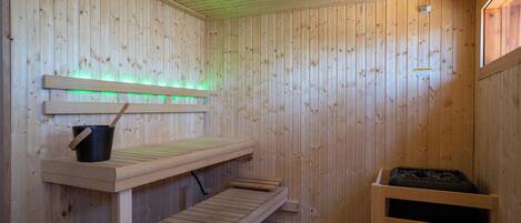 A sauna room with wooden walls, a bench, a bucket, and a heater.