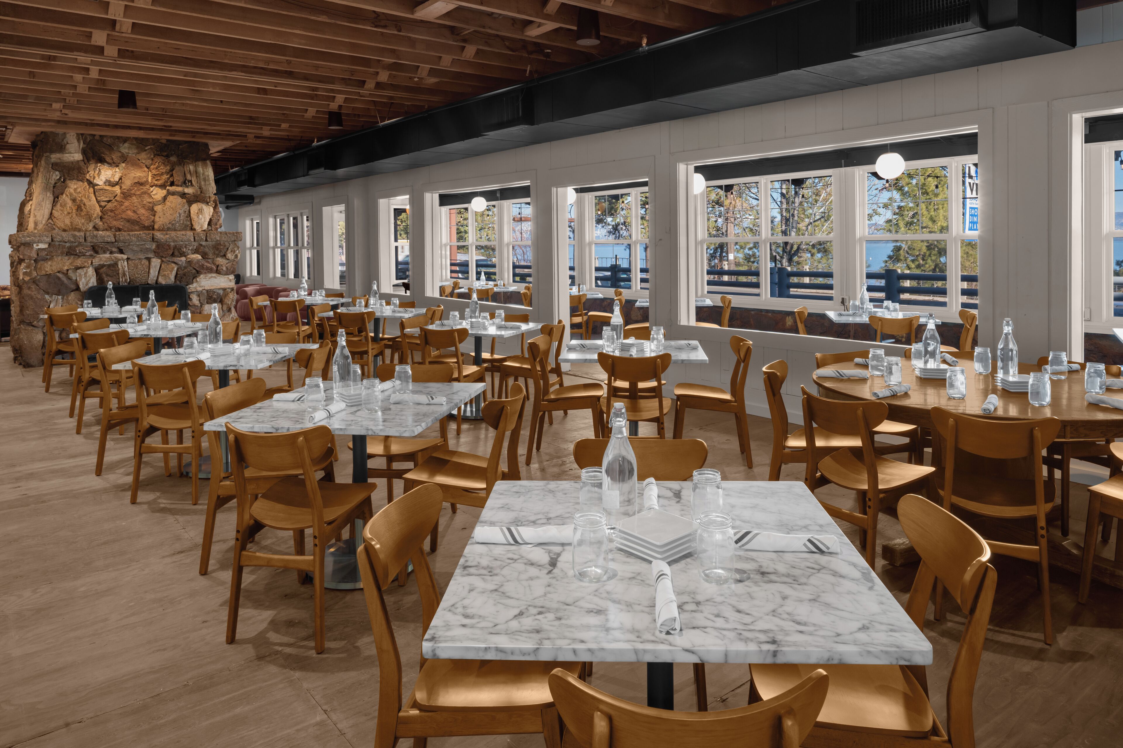 Un restaurante espacioso con mesas de mármol y sillas de madera, amplias ventanas con vistas al exterior y una chimenea de piedra.