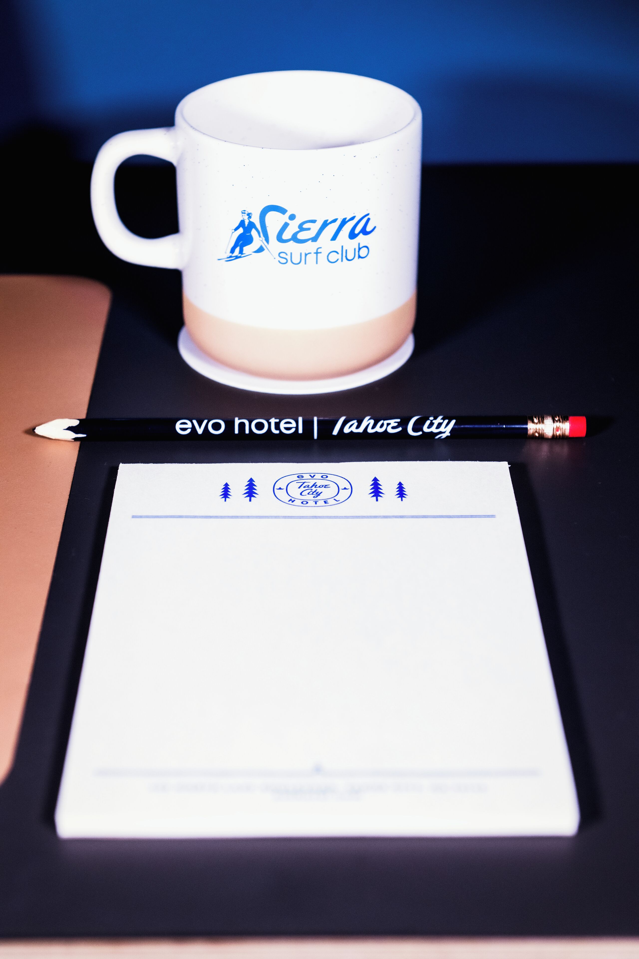 Une tasse blanche ornée d’un logo et d’un texte, un crayon noir et une feuille de papier contenant du texte et des icônes, le tout posé sur une surface sombre.