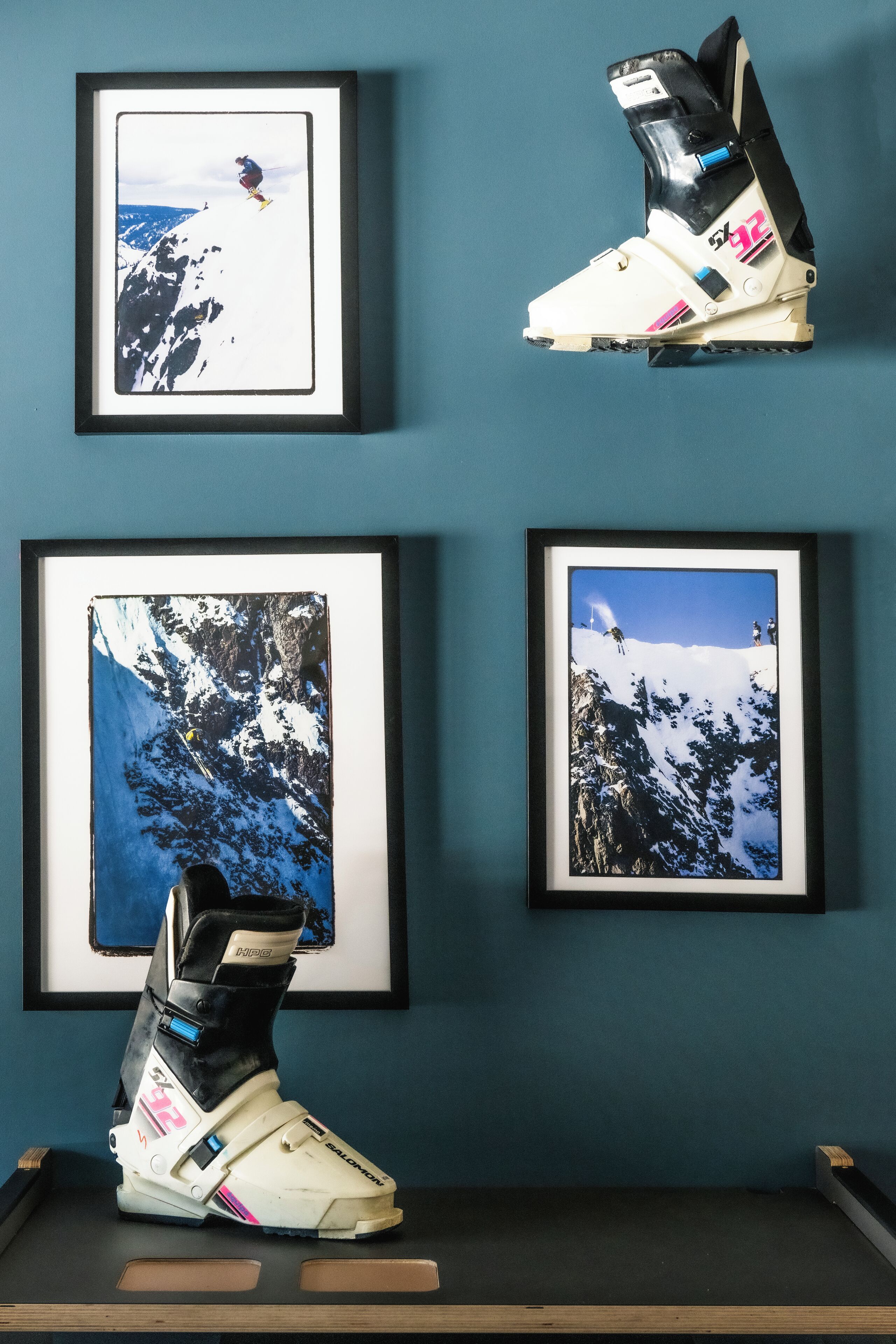 A pair of snow skis on a shelf with three framed photographs of snowy mountain landscapes on the wall behind them.