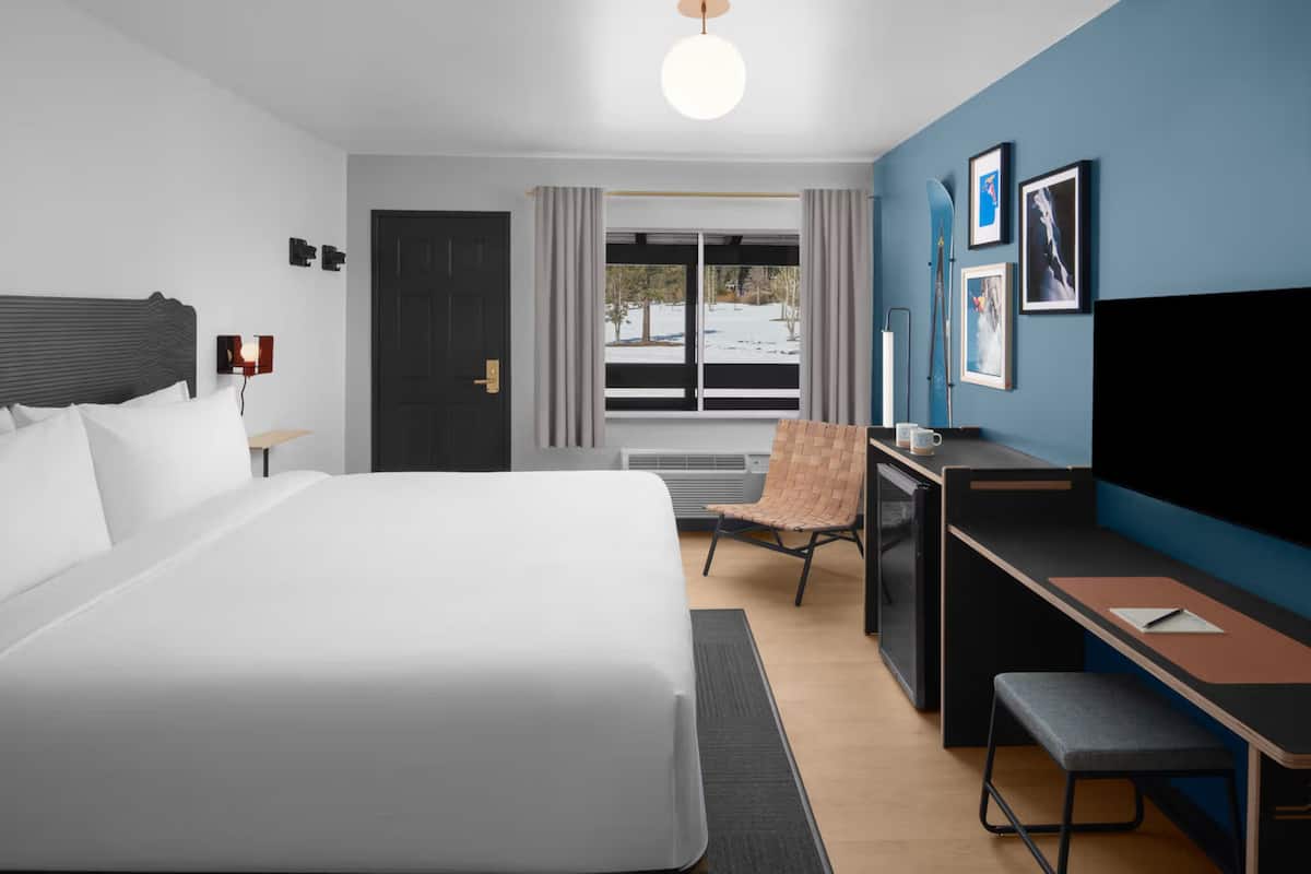 A modern hotel room with a large bed, a desk, a chair, a television, and a window with a view of trees and a building.