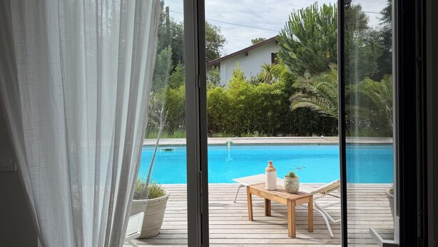 Une porte vitrée donne sur une terrasse en bois, où se trouvent une piscine, du mobilier d’extérieur et de la verdure.