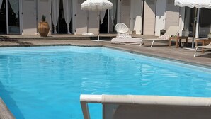 Une piscine entourée d’une terrasse en bois et de murs blancs, avec des chaises longues et des parasols.