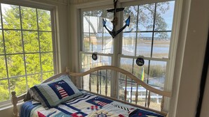 Een bed met een quilt in nautisch thema, een houten hoofdbord en uitzicht op een meer door grote ramen.