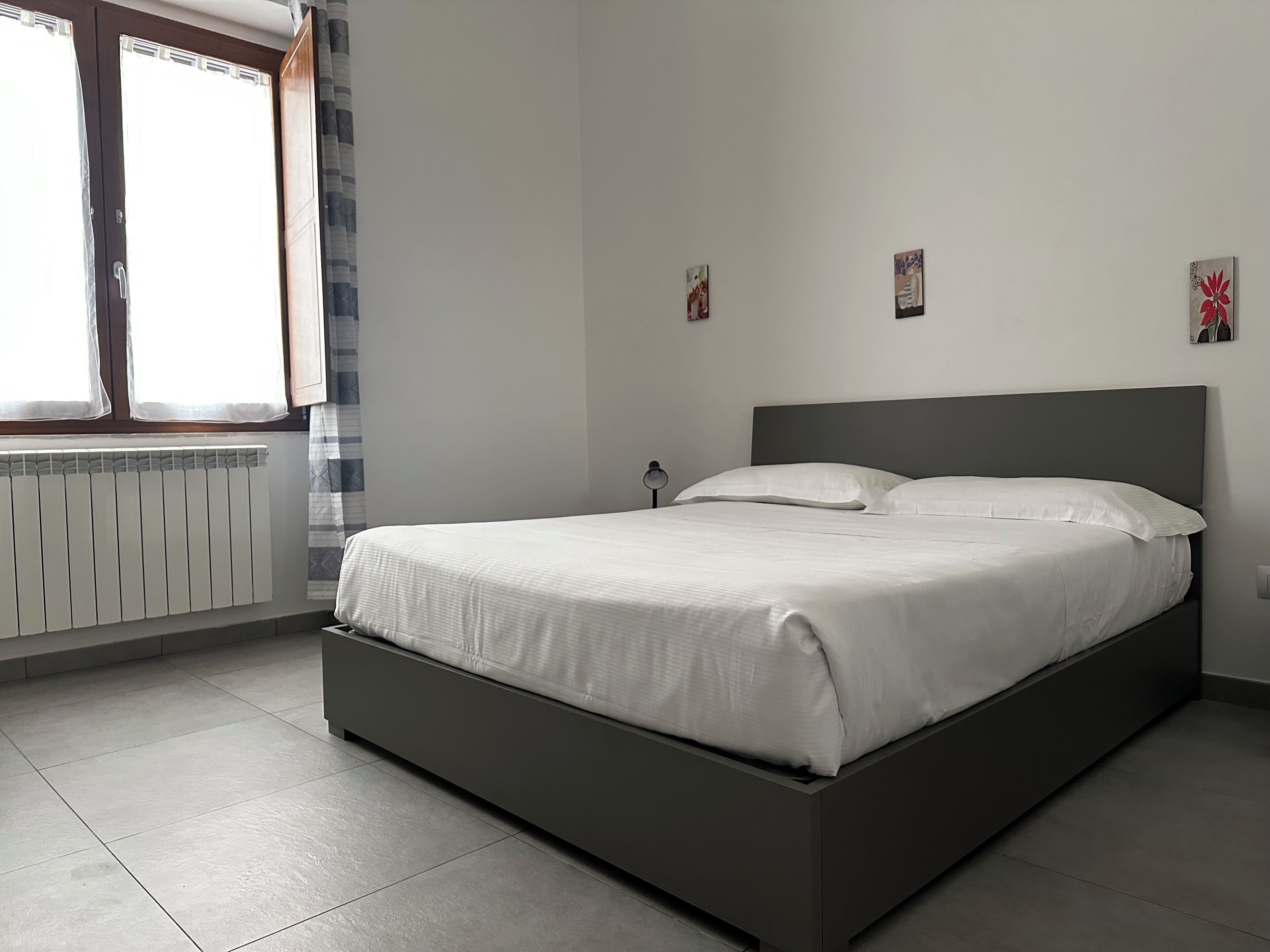 A bedroom with a bed, a radiator, a window with curtains, and a wall with three small pictures.