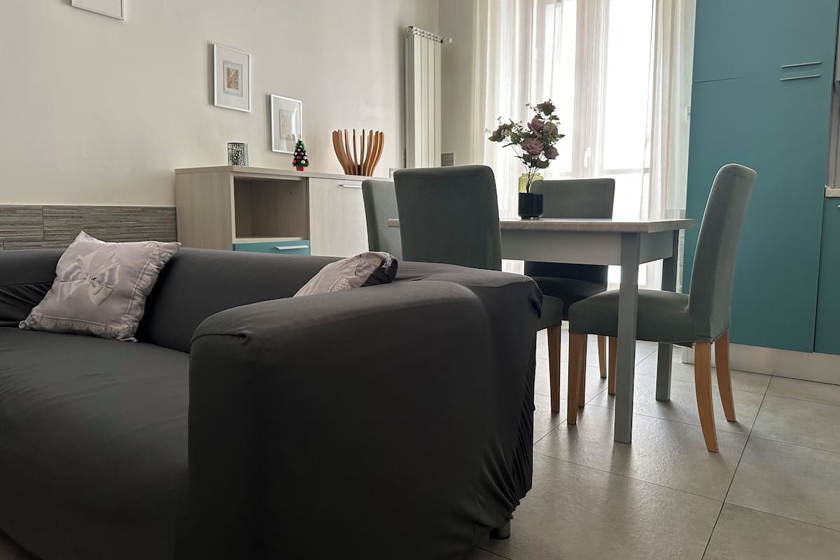 A modern living room with a grey sofa, a dining table with chairs, and a teal cabinet.