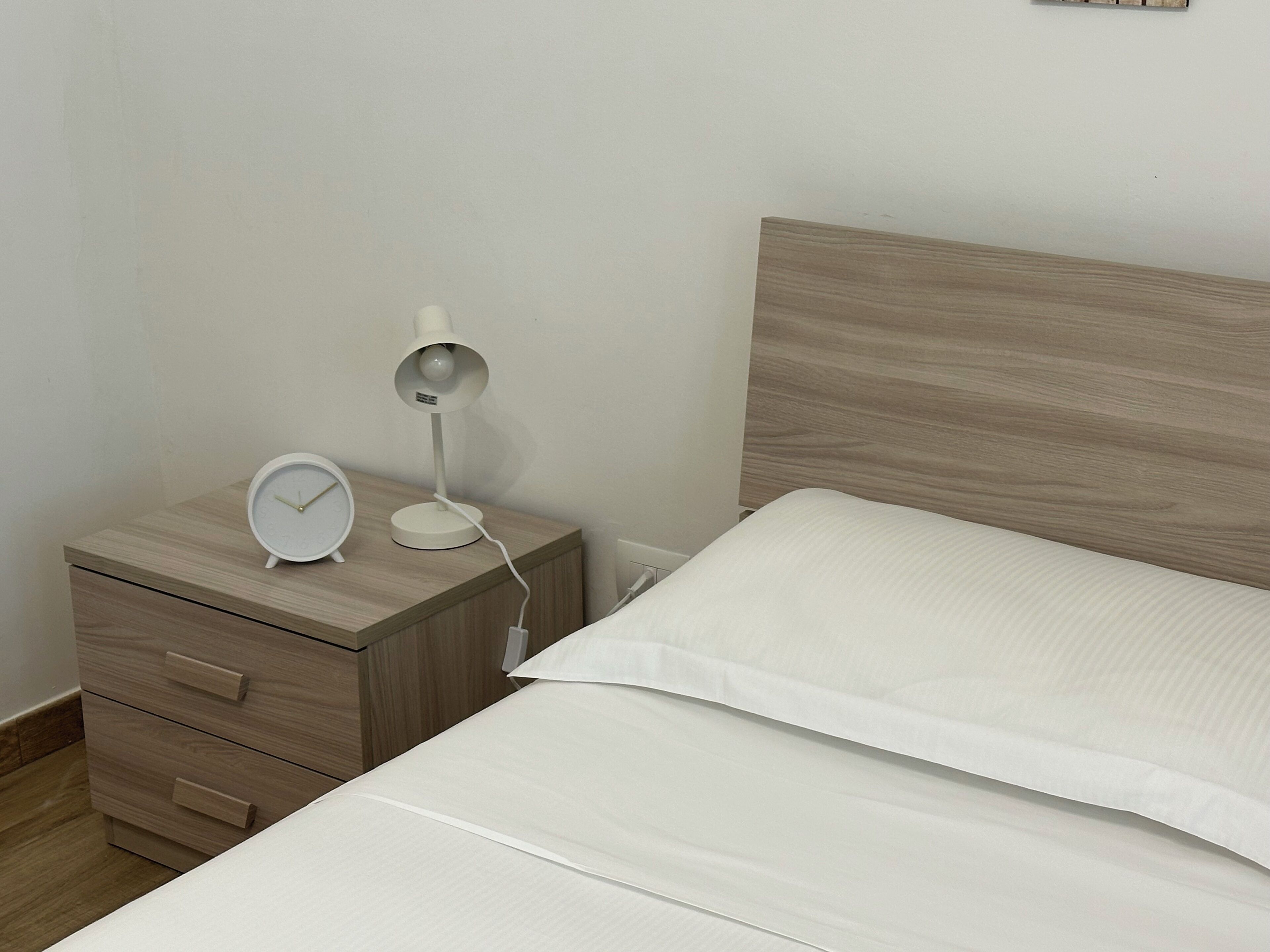 A neatly made bed with white linens, a wooden bedside table with a white alarm clock and a lamp, and a wooden headboard.