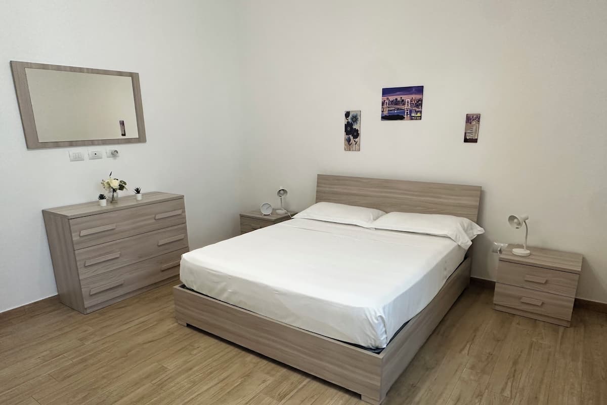 A bedroom with a bed, a dresser, a nightstand, a bedside table, a mirror, and a wall-mounted picture.