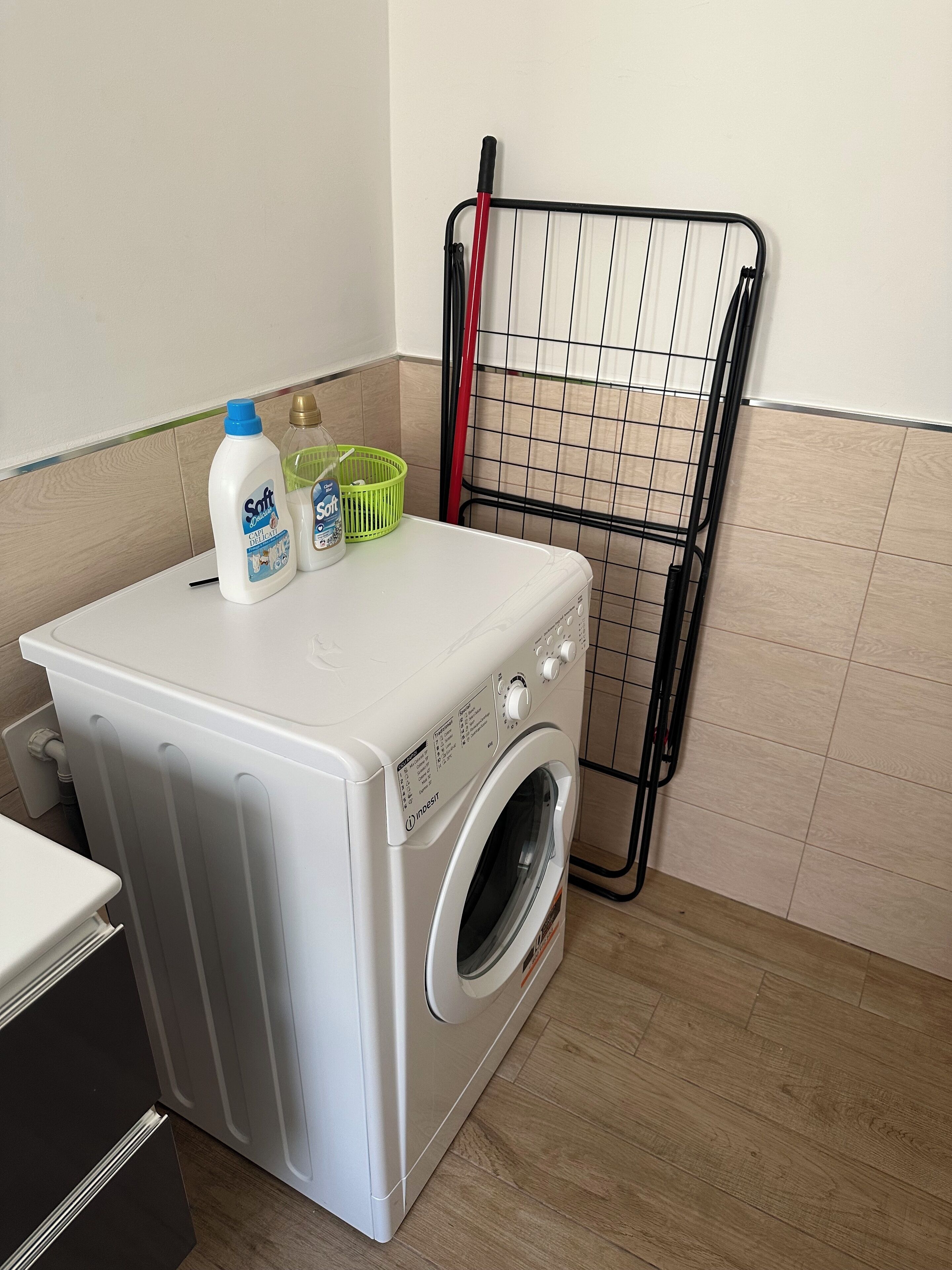 Una lavadora blanca y compacta con un panel de control, una cesta de ropa verde y dos botellas de detergente encima.