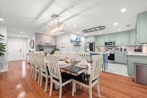 A modern kitchen with light green cabinets, stainless steel appliances, and a dining area with a wooden table and white chairs.