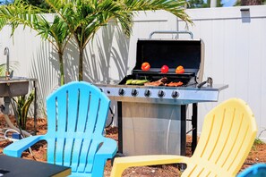 Un barbecue avec des légumes et de la viande, deux chaises Adirondack aux couleurs vives et, au fond, une clôture blanche.