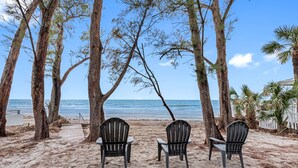 Una playa con tres sillas Adirondack mirando al océano, rodeada de árboles altos y una valla blanca.