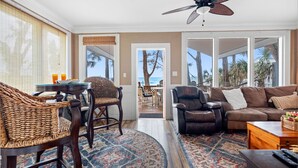 A spacious living room with a large sofa, a dining area with wicker chairs, a ceiling fan, and a view of the ocean through large windows. - Coastal Escape in Indian Rocks | Breathtaking Waterfront Views from the Deck (Indian Rocks Beach)