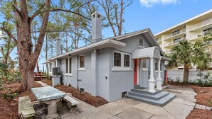 Una casa pequeña de un solo piso, con fachada gris, puerta roja y un porche con banco.