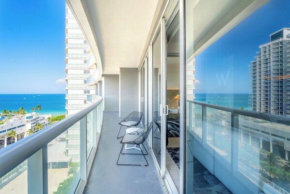 A balcony with a view of the ocean and buildings. There are two chairs and a table on the balcony.