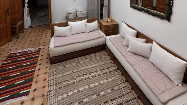 Ein geräumiges Zimmer mit hoher Decke und Oberlicht, ausgestattet mit einem großen Bett mit weißer Bettwäsche, einem Nachttisch aus Holz, einem Spiegel und einem gemusterten Teppich.