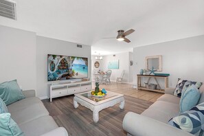 Un salon doté d’un grand téléviseur, d’un ventilateur de plafond et d’un coin repas avec une table et des chaises.