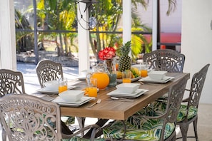 Une table à manger dressée pour six personnes, offrant une vue sur un paysage tropical, et entourée de chaises en métal aux motifs décoratifs.