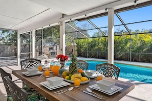 Un espace de restauration en plein air avec vue sur la piscine, comprenant une table dressée pour deux personnes, avec assiettes, verres et un panier de fruits.