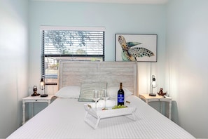Une chambre à coucher avec un lit, des tables de chevet, une fenêtre avec des stores, un tableau encadré au mur et un plateau avec des verres à vin et des raisins sur le lit.