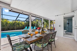 An outdoor dining area with a pool view, surrounded by tropical plants and equipped with a table, chairs, and a glass enclosure. - Screened Heated Pool, 15 Min to Beach | 4BR Sleeps 10, Largo (Largo)