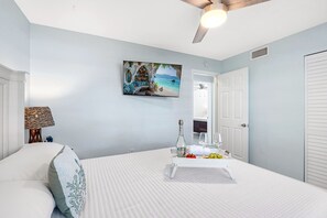 Une chambre à coucher avec un lit, une table de chevet sur laquelle se trouvent une bouteille et des verres, un tableau représentant une scène de plage et un ventilateur de plafond.