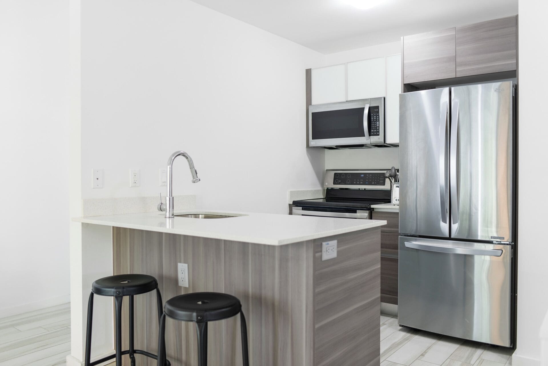 A modern kitchen with a stainless steel refrigerator, a microwave, a stove, and a sink. There are two black stools at the counter.