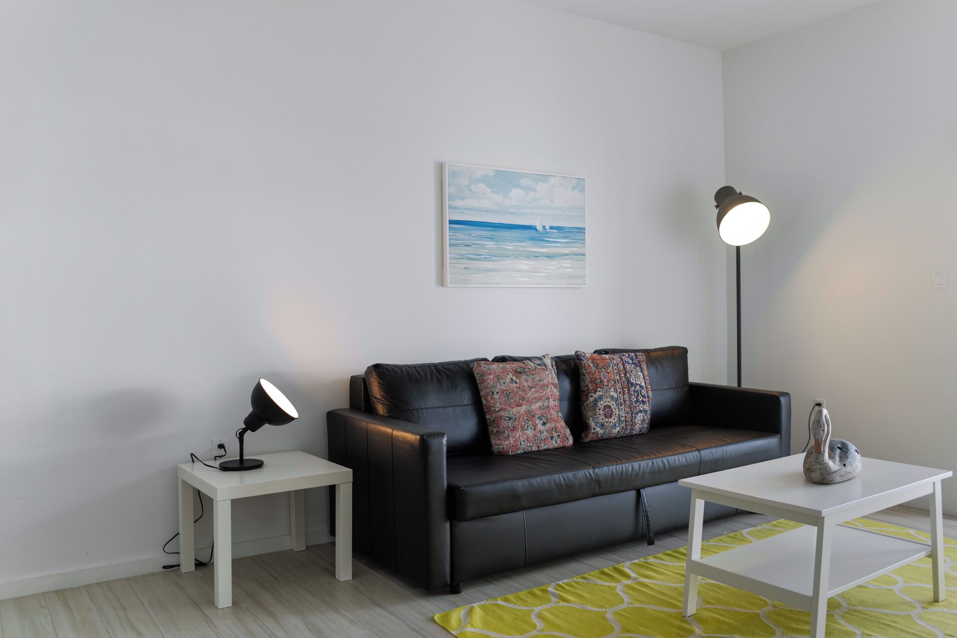 A modern living room with a black leather sofa, patterned cushions, a white coffee table, a floor lamp, and a painting of a seascape.