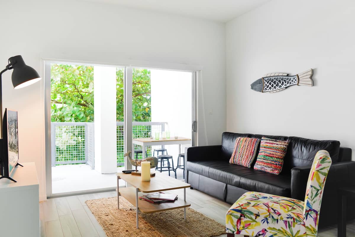 Un salon moderne avec un canapé en cuir noir, des coussins colorés, une table basse en bois et une grande fenêtre donnant sur un paysage verdoyant.