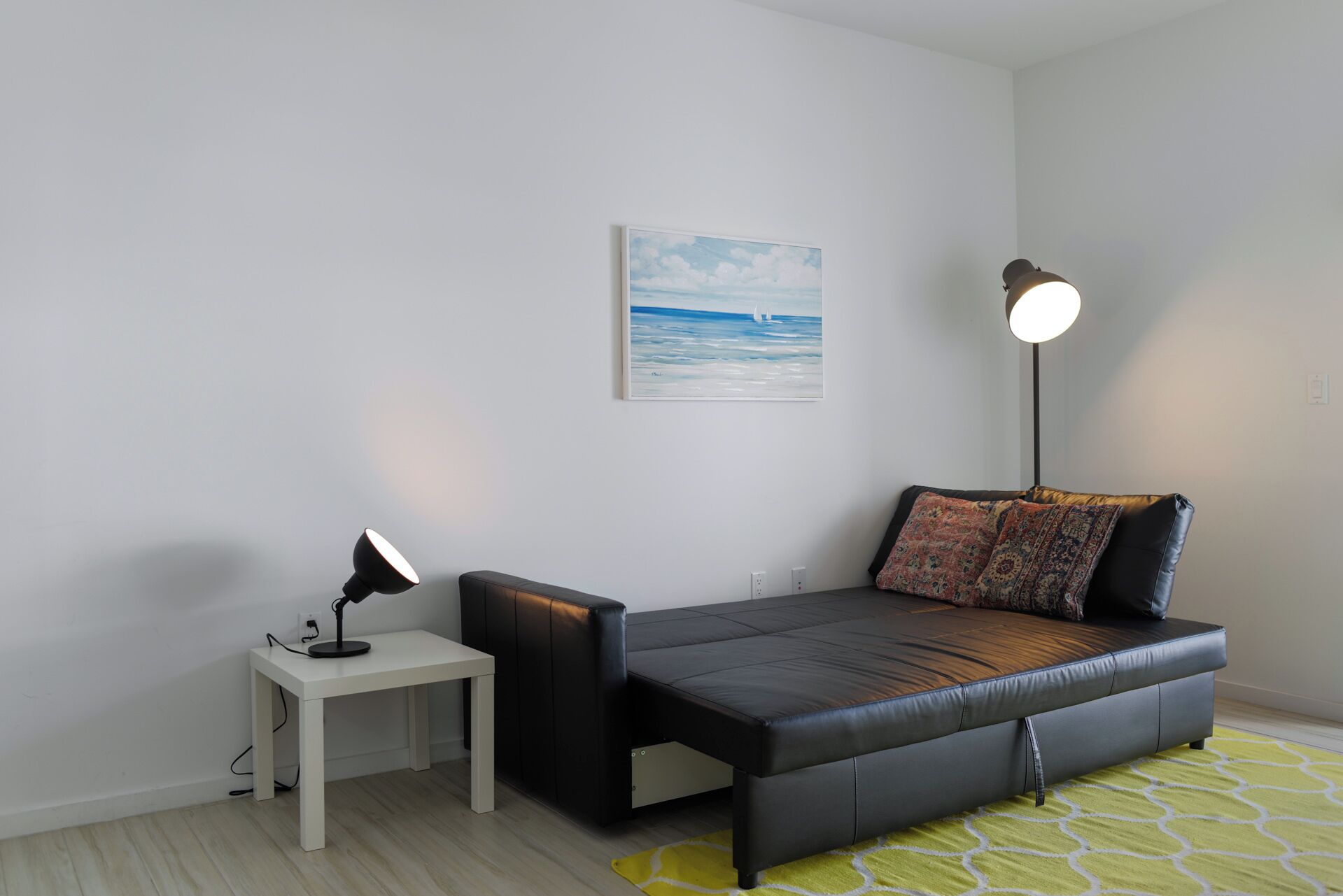 Una sala de estar moderna con un sofá de cuero negro, una mesita auxiliar blanca, una lámpara de pie y un cuadro de un velero en la pared.