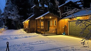 Eine Holzhütte mit schneebedecktem Dach, einer sichtbaren Garagentür und einem Holzstapel davor.