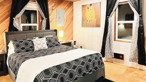 Ein Schlafzimmer mit Holzbalkendecke, einem Bett mit geometrisch gemustertem Überwurf, schwarz-weißen Vorhängen und einem Bild an der Wand.
