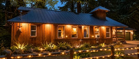 A wooden cabin with a metal roof, surrounded by a gravel path and a wooden fence with string lights.